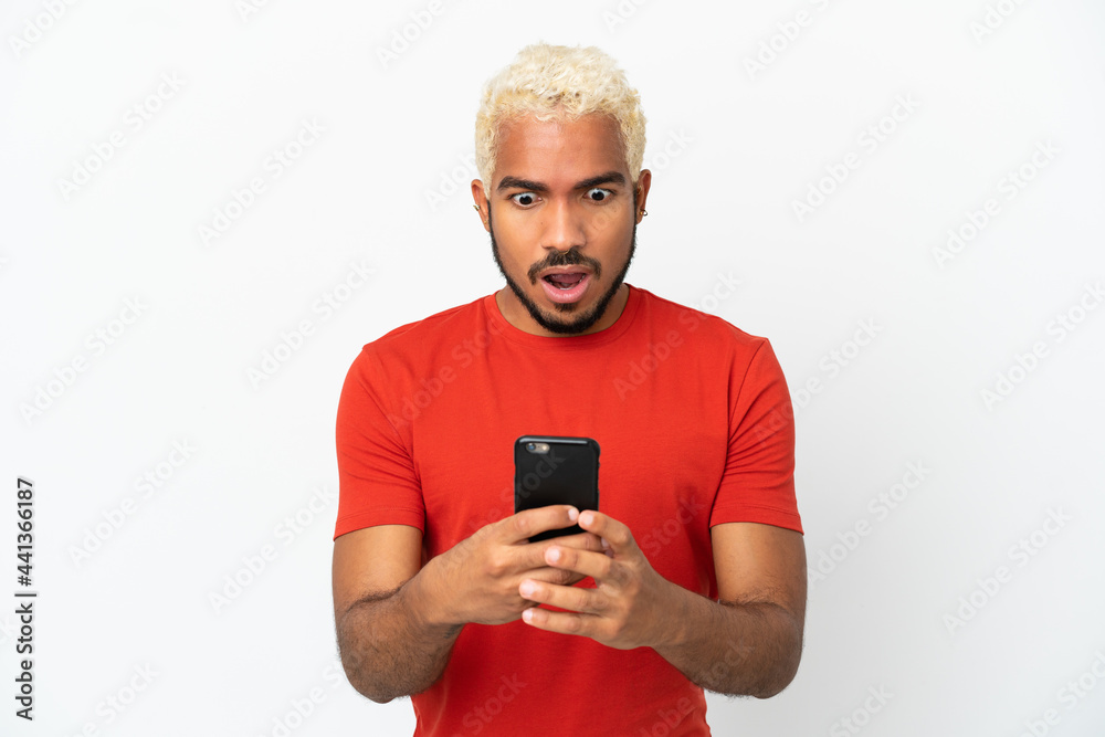 Young Colombian handsome man isolated on white background looking at the camera while using the mobile with surprised expression Young Colombian handsome man isolated on white background looking at the camera while using the mobile with surprised expression