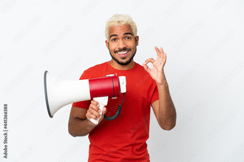 Young Colombian handsome man isolated on white background holding a megaphone and showing ok sign with fingers Young Colombian handsome man isolated on white background holding a megaphone and showing ok sign with fingers