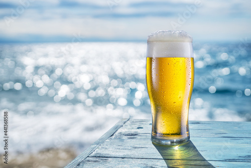 Cooled glass of beer with condensation drops on the blue wooden table. Blurred sea is at the background. photo