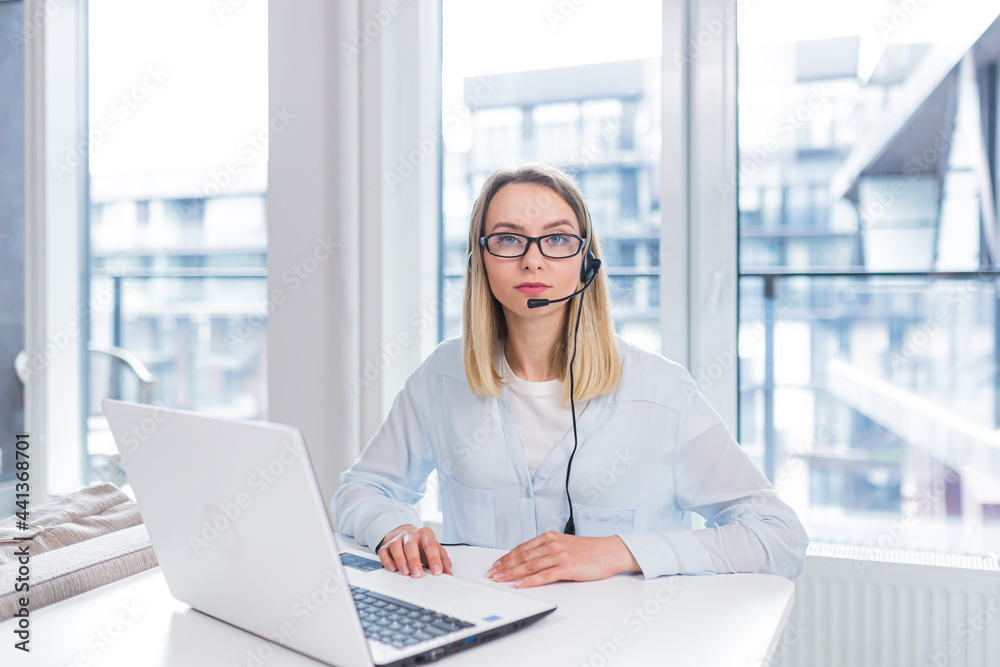 Young blonde woman with a headset and glasses communicates with customers at a distance in front of a laptop or computer screen in the office of an online consumer support call center. conference help Young blonde woman with a headset and glasses communicates with customers at a distance in front of a laptop or computer screen in the office of an online consumer support call center. conference help