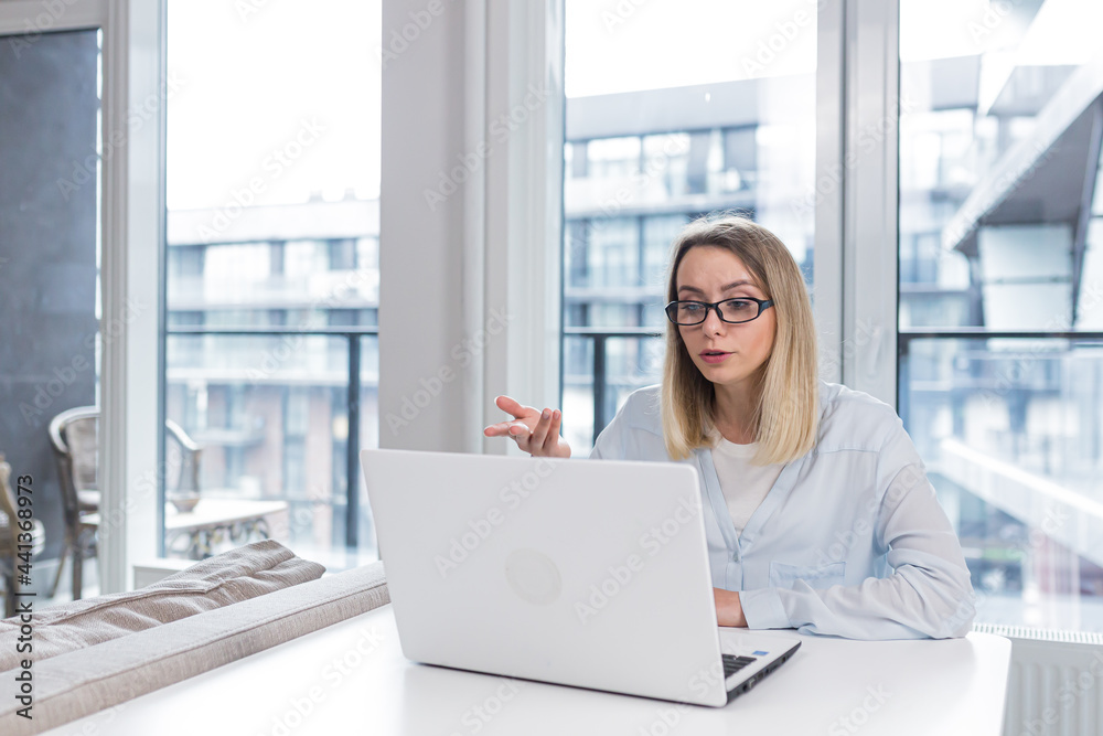 Caucasian happy young freelancer woman sitting at table and talking online on webcam remotely. Female student tells and communicates video call with friends or family indoors. Home or modern office Caucasian happy young freelancer woman sitting at table and talking online on webcam remotely. Female student tells and communicates video call with friends or family indoors. Home or modern office