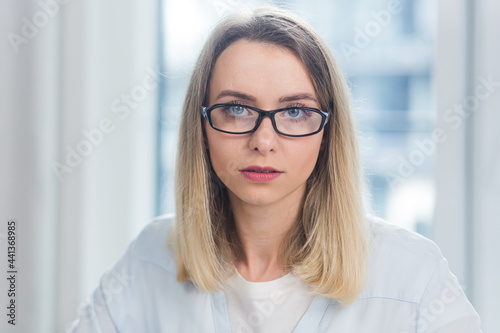 Close-up portrait of a young adult blonde business woman looking at the camera indoors at office or home on a blue window background. Face Confident female Manager in Formal Wear indoors
