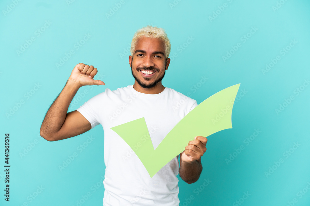 Young Colombian handsome man isolated on blue background holding a check icon with proud gesture Young Colombian handsome man isolated on blue background holding a check icon with proud gesture