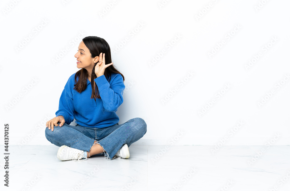Young mixed race woman sitting on the floor isolated on white background listening to something by putting hand on the ear