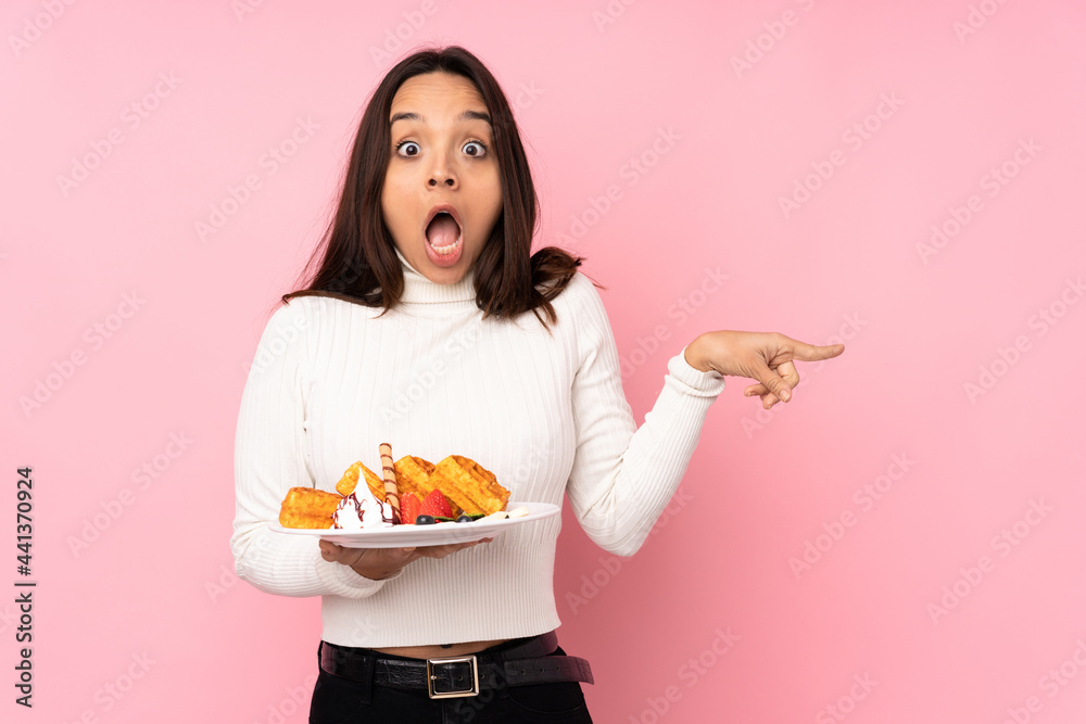 Young brunette woman holding waffles over isolated pink background surprised and pointing side Young brunette woman holding waffles over isolated pink background surprised and pointing side
