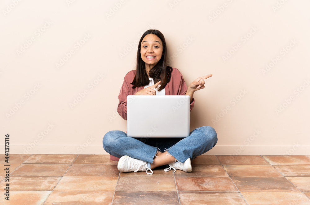 Young mixed race woman with a laptop sitting on the floor frightened and pointing to the side Young mixed race woman with a laptop sitting on the floor frightened and pointing to the side