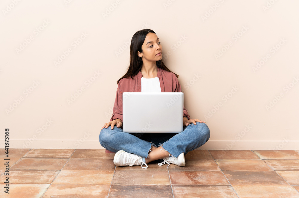 Young mixed race woman with a laptop sitting on the floor in back position and looking side Young mixed race woman with a laptop sitting on the floor in back position and looking side