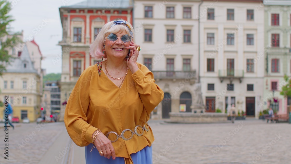 Excited elderly old tourist woman talking on mobile phone while walking on city street of Lviv, Ukraine. Senior stylish mature granny enjoying smartphone conversation and traveling. Urban concept