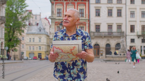 Elderly tourist stylish grandfather getting lost in big city trying to find route. Excited senior traveler man walking along street, looking for way using paper map in old town. Summer vacation trip