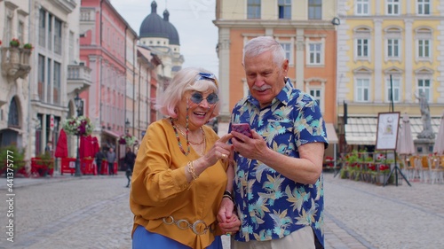 Senior old stylish tourists man and woman looking smartphone display sincerely rejoicing win, receiving message with good news, success luck. Elderly grandmother, grandfather enjoying conversation