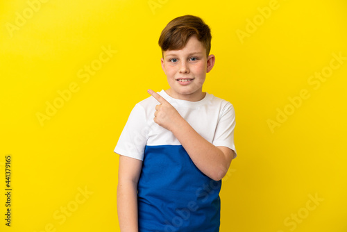 Little redhead boy isolated on yellow background pointing to the side to present a product