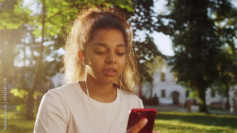 Mixed race young woman smiling and laughing using phone for video chat online with friends and blogging for social media while sitting on grass at the street. Mixed race young woman smiling and laughing using phone for video chat online with friends and blogging for social media while sitting on grass at the street.