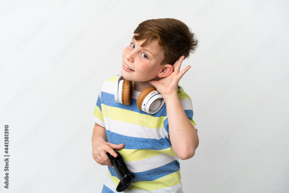 Little redhead boy holding a game pad isolated on white background listening to something by putting hand on the ear Little redhead boy holding a game pad isolated on white background listening to something by putting hand on the ear