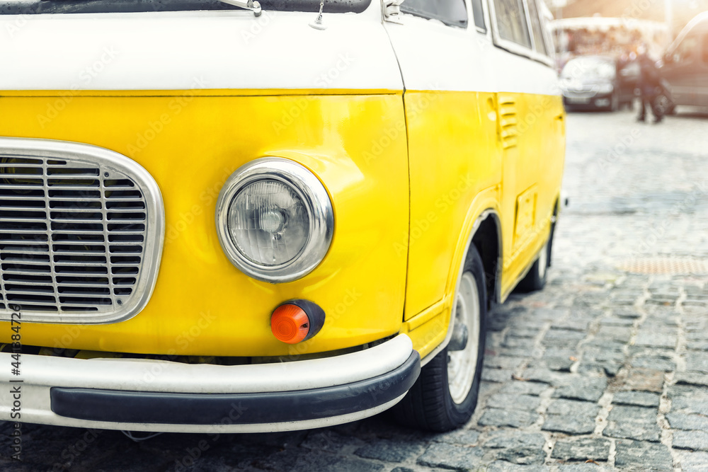 Close-up detail front view of headlight part old vintage bright yellow retro mini bus car van parked in european city center on cobble stone paved road. Fun vehicle for snack delivery sale journey