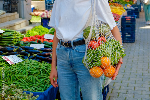 Plant based diet concept. Young woman at farmers market with reusable eco friendly net bag full of groceries. Conscious shopping for organic fruits and vegetables. Close up, copy space, background. photo