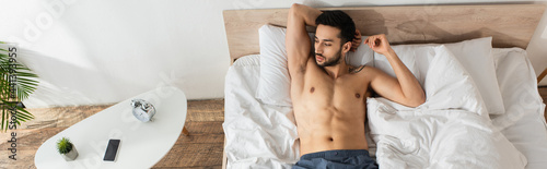 Top view of shirtless man lying near alarm clock and smartphone on bedside table, banner