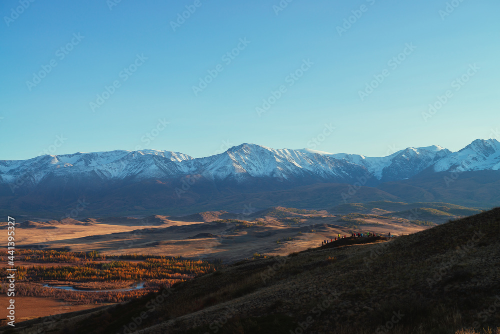 Awesome autumn landscape with tourists crowd on hill with view to red valley and great snow-covered mountain range in red sunset sunshine. Spectacular sunny view to snow mountains and autumn valley.