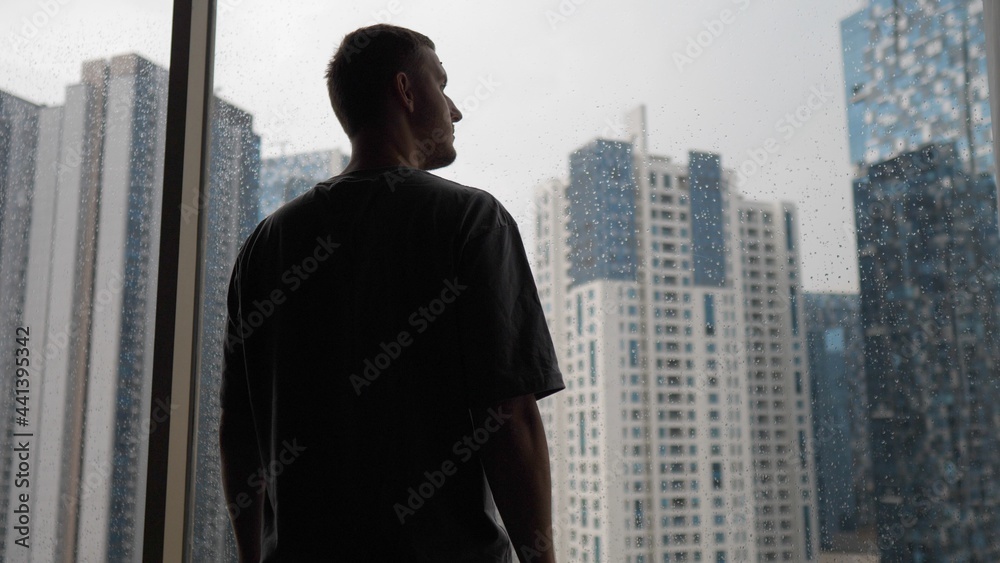 Loneliness in a big city. Back view of a young man looking outside his window on the skyscrapers