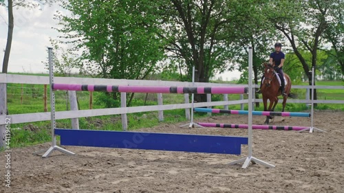 Female Jockey on a horse running and leaping over fences. Equestrian events concept. Beautiful brown horse jumping over obstacles. Competitive woman riding a Horse. Lush green trees in the background.