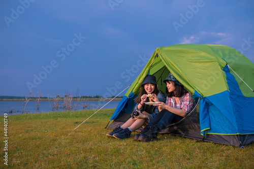 group of Asian friends tourist drinking and playing guitar together with happiness in Summer while having camping near lake at sunset