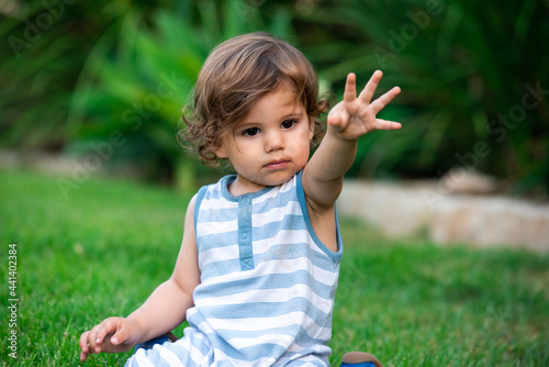 A 1.5 years old boy is sitting on a green grass in a garden and raising his hand while playing.
