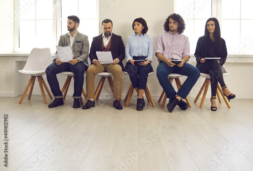 Candidates with documents and resumes in their hands sit on chairs in the lobby waiting for an interview. Different men and women compete for the same position. Staff employment concept.