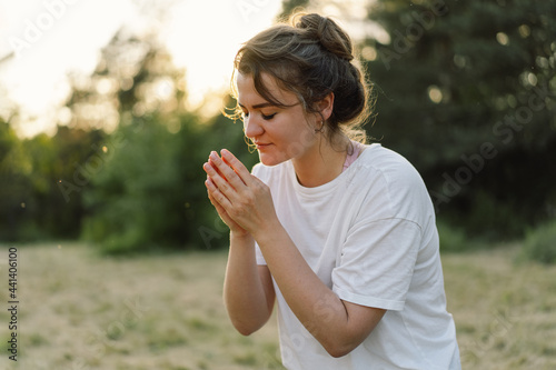 Woman closed her eyes, praying in a field during beautiful sunset. Hands folded in prayer concept for faith, spirituality and religion.
