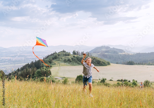 Smiling teenager boy with flying colorful kite on the high grass meadow in the mountain fields. Happy childhood moments or outdoor time spending concept image. #441412144