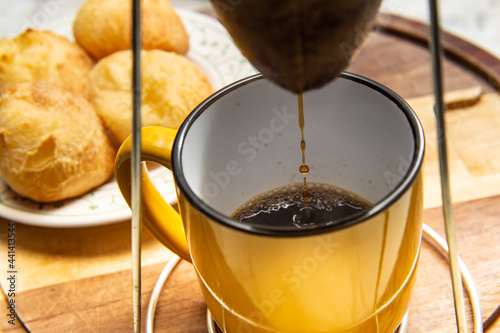 Café sendo coado no coador com uma caneca amarela abaixo com um prato de pão de queijo ao fundo. photo