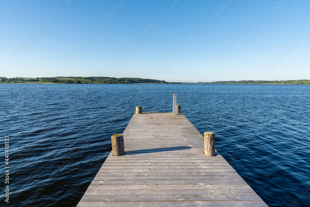wooden boardwalk leading out into the dark blue waters of a fjord under a cloudless blue sky