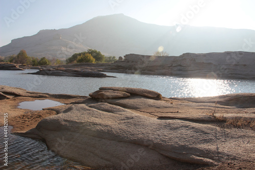 A small lake in the volcanic low rocks in the Bektau Ata tract, the sun is reflected in the water, a sunbeam, bushes and a mountain in the distance, early morning, summer, sunny photo