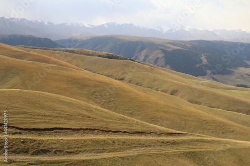Top view of the soft slopes of hills and mountains on the Assy plateau, in the distance a snow-covered ridge in a light fog, a sky with gentle clouds, summer, sunny