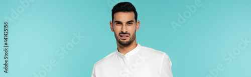 Confused arabian man in white shirt looking at camera isolated on blue, banner