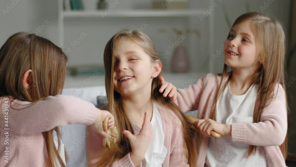 Three funny cute little girls sisters best friends triplets twins same children sitting at home couch two kids combing long smooth hair of one schoolgirl using combs brushing make hairstyle coiffure Three funny cute little girls sisters best friends triplets twins same children sitting at home couch two kids combing long smooth hair of one schoolgirl using combs brushing make hairstyle coiffure