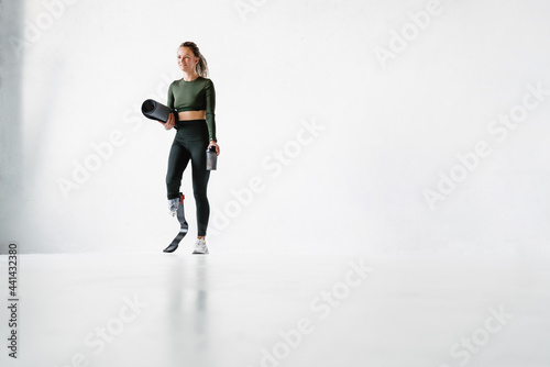 Young sportswoman with prosthesis holding fitness mat and water bottle