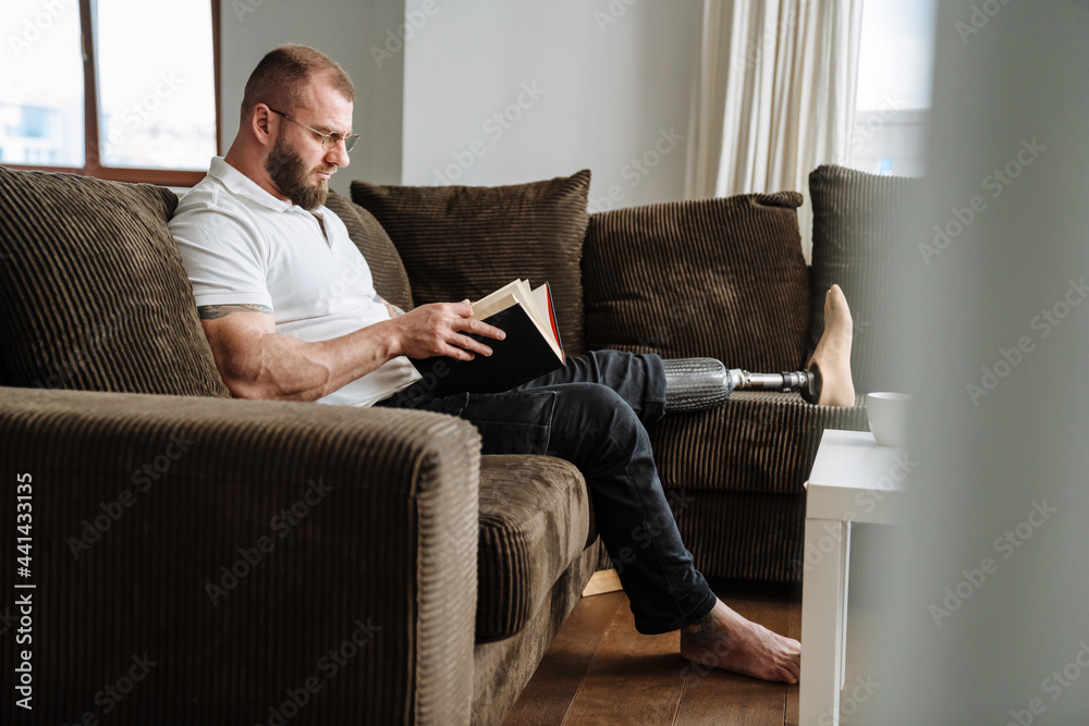 White man with prosthesis reading book while siting on couch at home White man with prosthesis reading book while siting on couch at home
