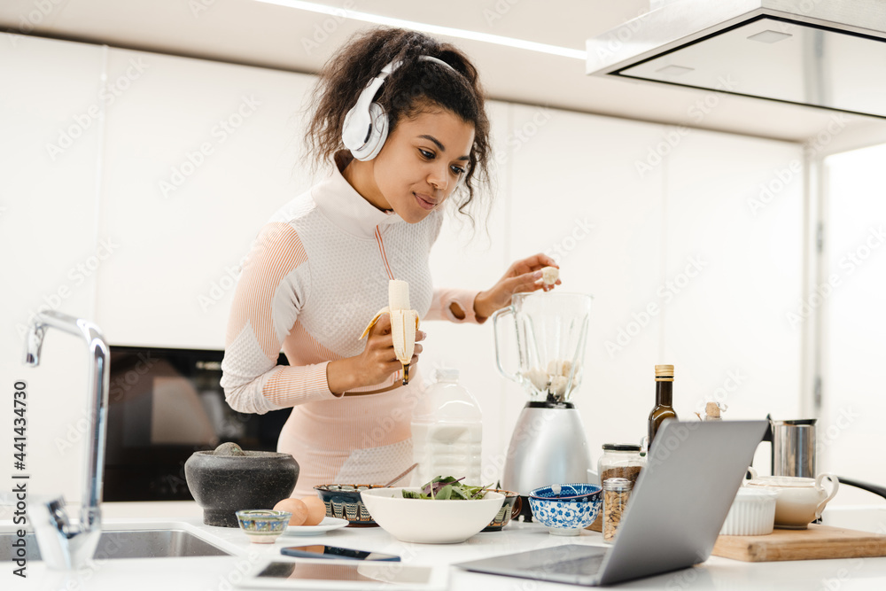 Black woman in headphones using laptop while making smoothie at home