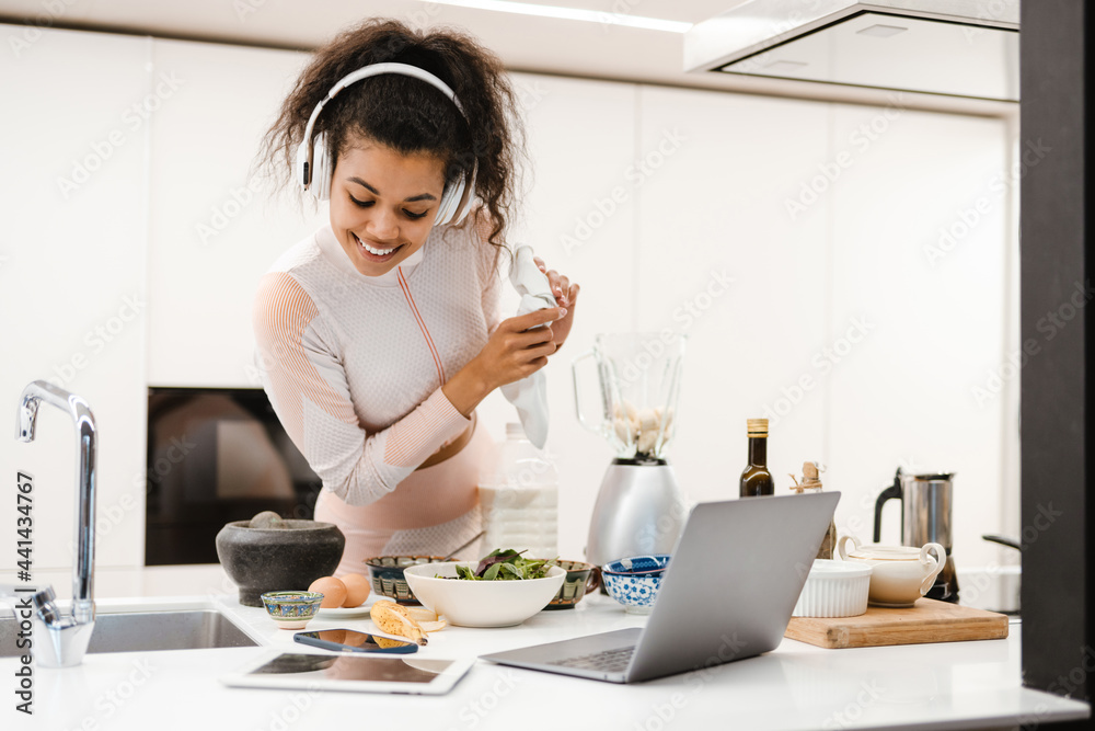 Black woman in headphones using laptop while making smoothie at home Black woman in headphones using laptop while making smoothie at home