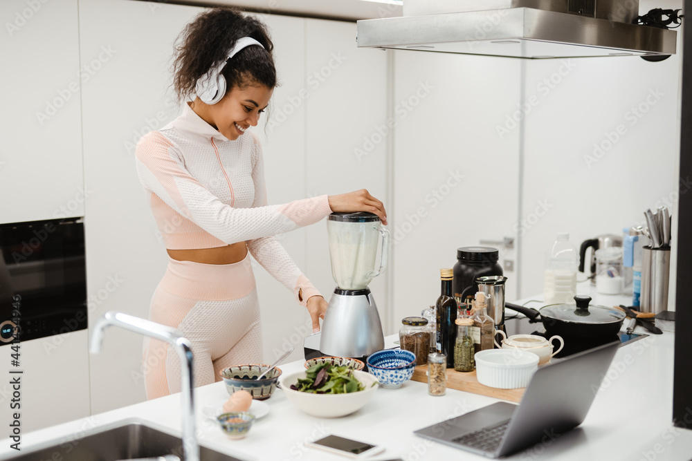 Black woman in headphones using laptop while making smoothie at home Black woman in headphones using laptop while making smoothie at home