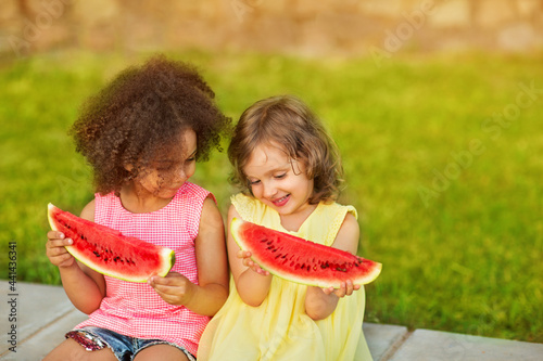 Funny Black girl and European girl are eating watermelon outdoors in the hot summer. Smiling children enjoy healthy food