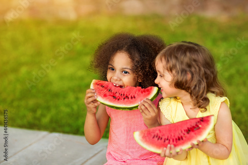 Funny Black and European girls are eating watermelon outdoors in the hot summer. Smiling children enjoy healthy food