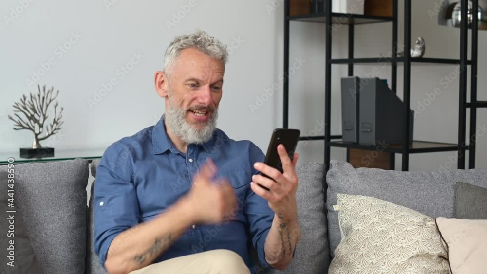 Happy middle aged man making video call on the smartphone, using mobile app for video conect sitting on the couch at home with phone in hands, looks at the camera waving and talking online