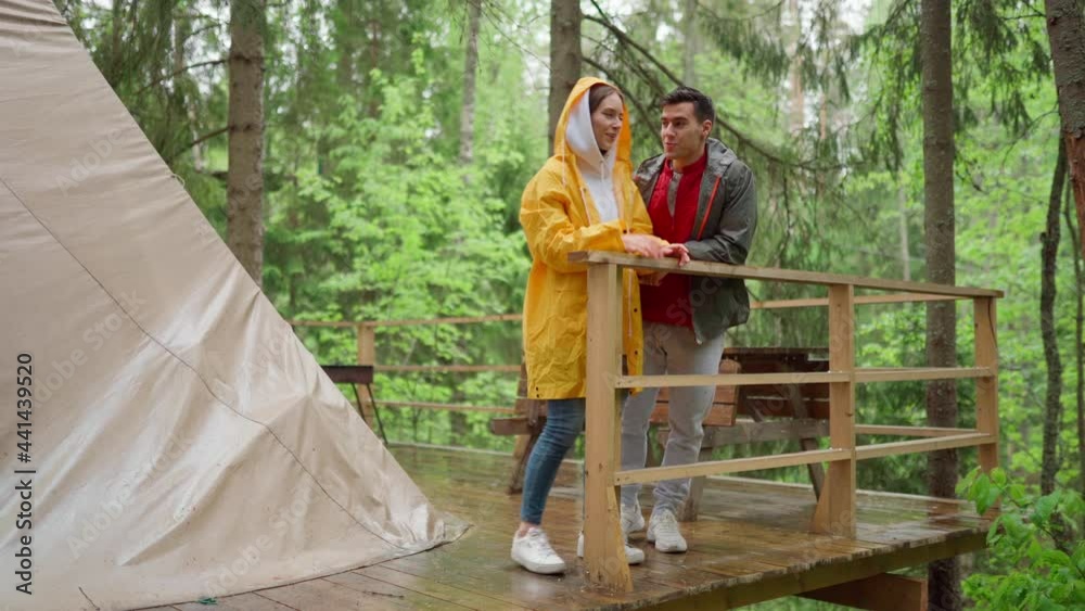Young female hiker standing on wooden porch next to tent in woodland and enjoying rain, boyfriend joining her. Couple of travelers talking cheerfully and admiring beautiful nature in forest Young female hiker standing on wooden porch next to tent in woodland and enjoying rain, boyfriend joining her. Couple of travelers talking cheerfully and admiring beautiful nature in forest