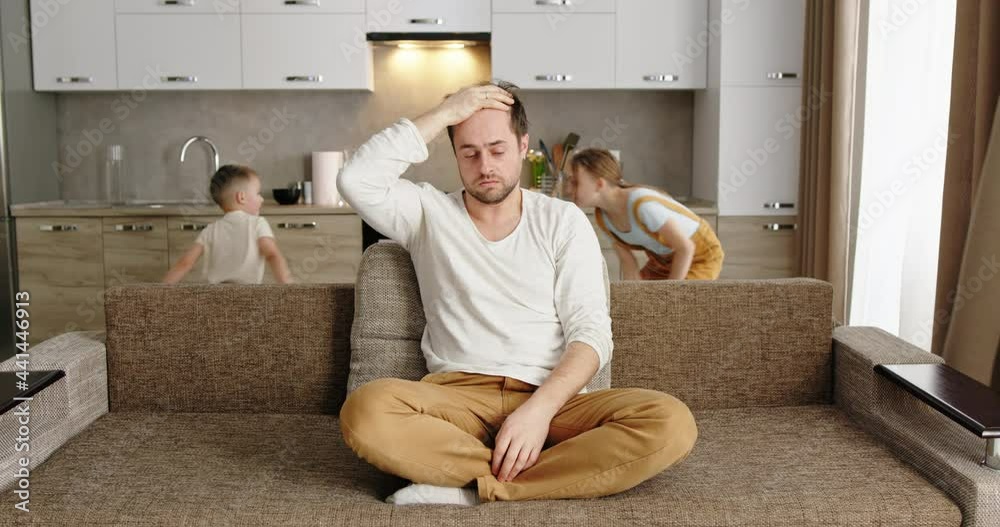 Father sitting on sofa at home exhausted frustrated tired holding his head headache 2 children running around the sofa shouting throwing pillows