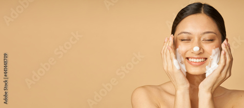 Website header of Studio portrait of pleased young woman smiling with eyes closed while applying gentle foam facial cleanser isolated over beige background photo