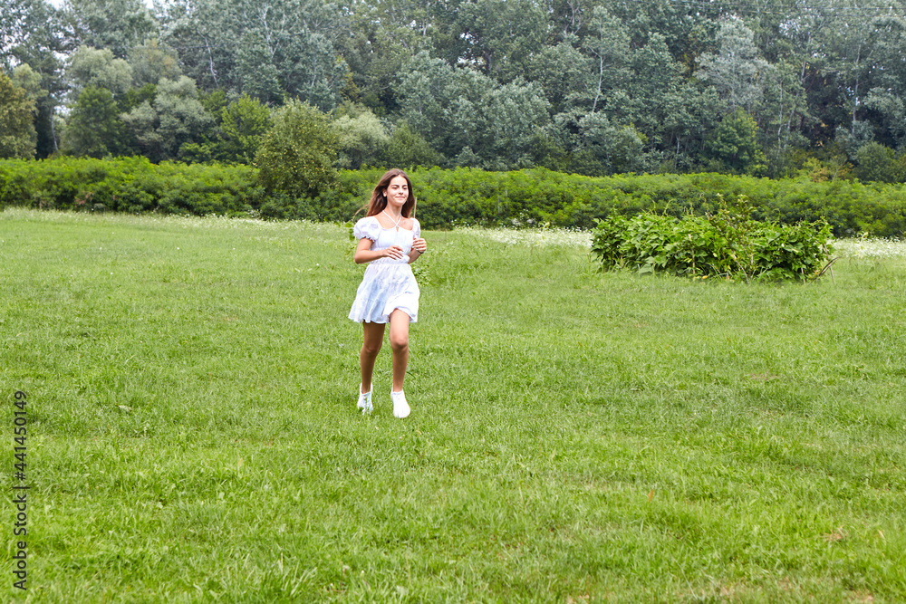 A young beautiful girl is running through a meadow with green grass in a white dress. The concept of joy for a summer day, happiness and youth
