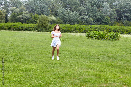 A young beautiful girl is running through a meadow with green grass in a white dress. The concept of joy for a summer day, happiness and youth