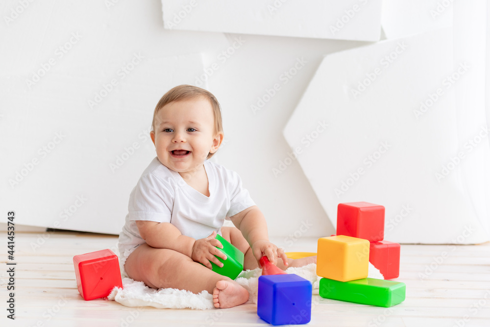 happy little baby six months old in a white t shirt and diapers playing at home on a Mat in a bright room with bright colored cubes happy little baby six months old in a white t shirt and diapers playing at home on a Mat in a bright room with bright colored cubes