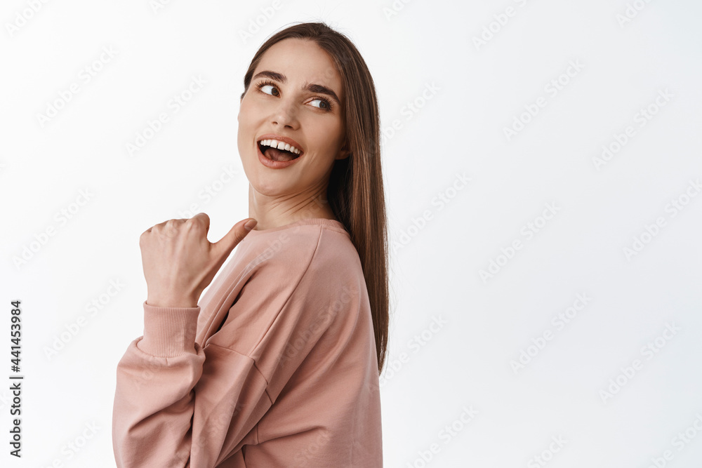 Fototapeta premium Portrait of cheerful beautiful girl pointing thumb and look behind her shoulder, laughing and smiling while looking and showing advertisement logo, white background