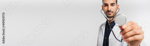Arabian doctor holding stethoscope on blurred foreground isolated on grey, banner